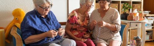 Trois femmes assises sur un canapé participent à une activité de crochet, entourées de pelotes et d’ouvrages.